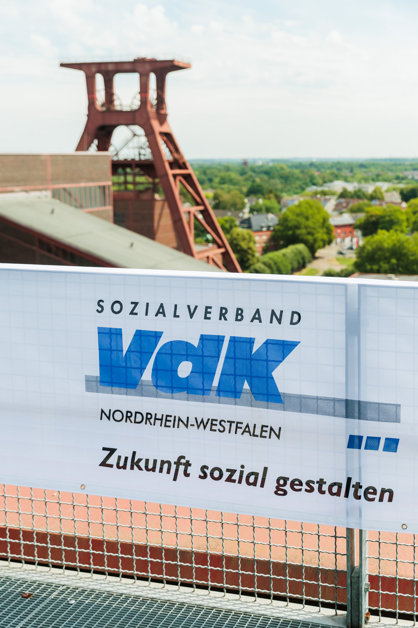 VdK-NRW-Banner an einem Zaun, dahinter der Förderturm der Zeche Zollverein und viele Baumwipfel.