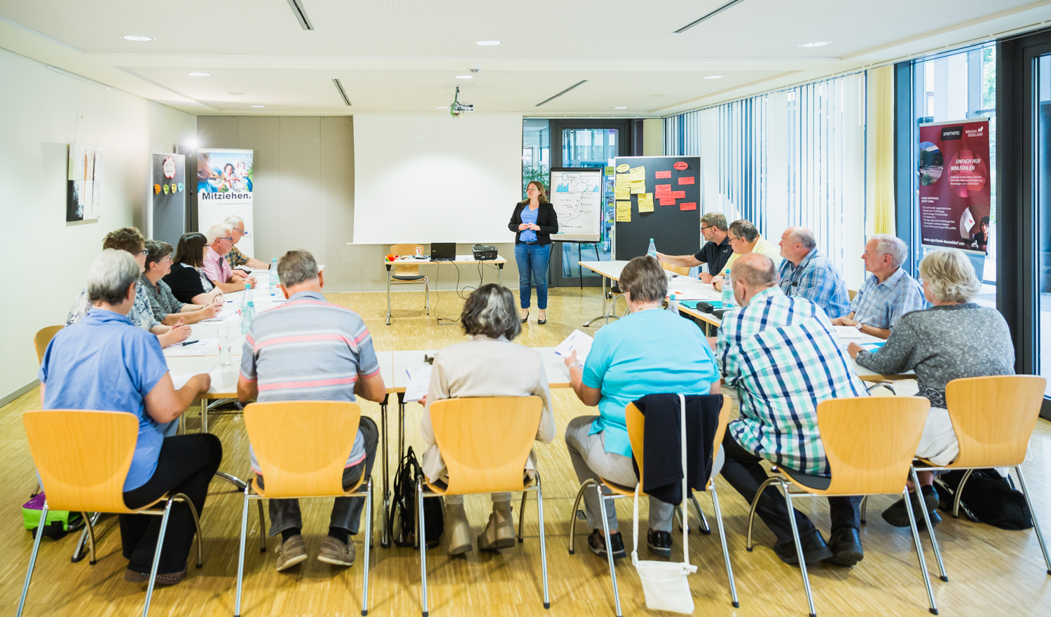 Menschen sitzen in einem Seminarraum an u-f&ouml;rmig aufgestellten Tischen. Vor Kopf steht eine Referentin vor einem White-Board.