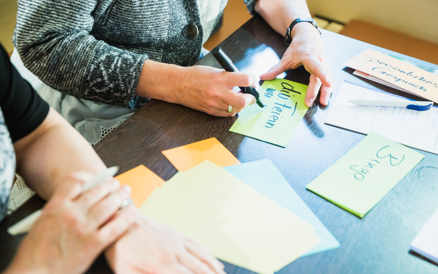 Man sieht von zwei Personen an einem Tisch nur die Arme und Hände. Auf dem Tisch bunte Karteikarten und Stifte.
