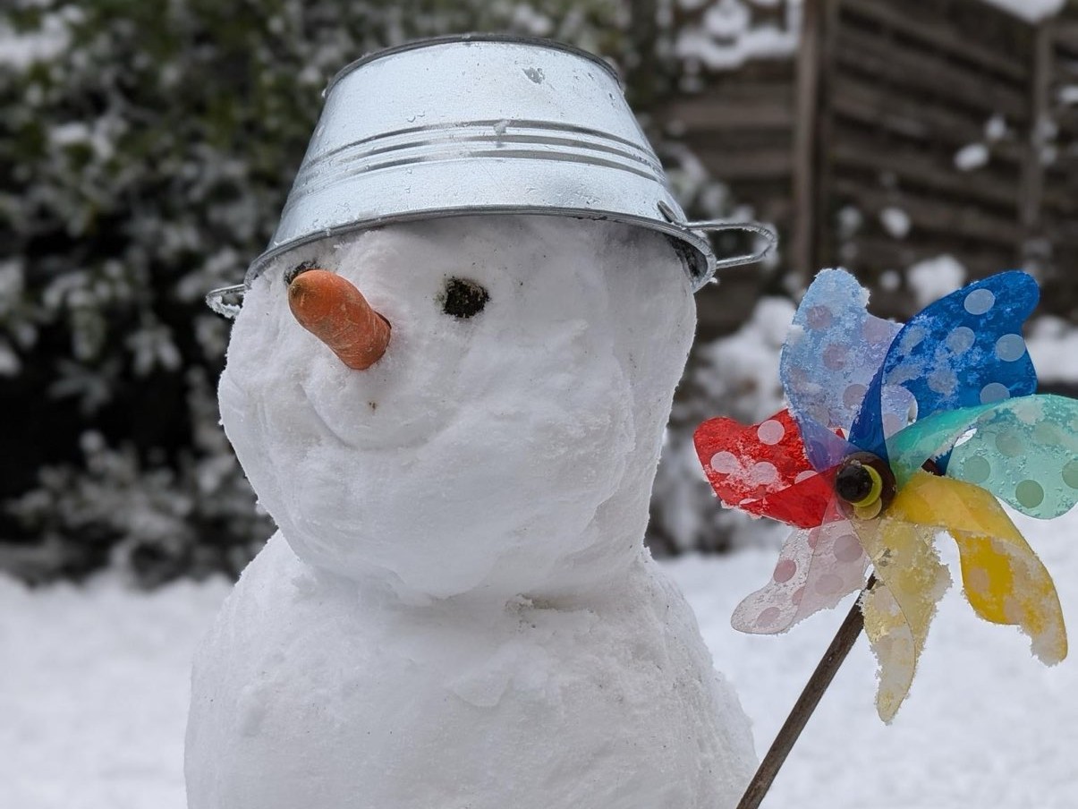 Ein Schneemann mit Möhrennase, Metallschüsselhut und buntem Windrad vor verschneiter Landschaft.