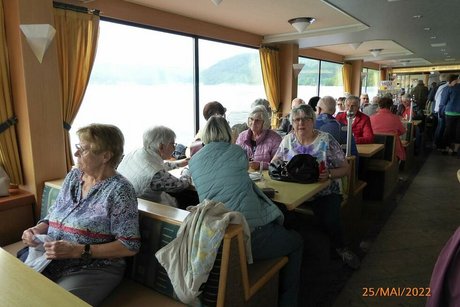 Eine Gruppe Menschen sitzt an Tischen an Deck des Fährschiffes.