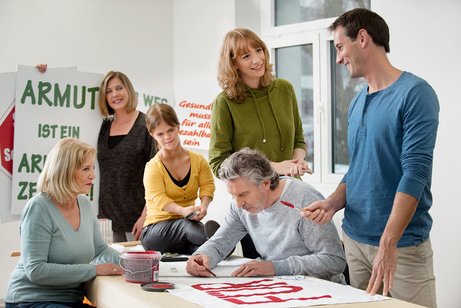 Eine Gruppe unterschiedlicher Menschen - Männer, Frauen, Alte und Junge, mit und ohne Behinderung, malt gemeinsam Demo-Plakate.