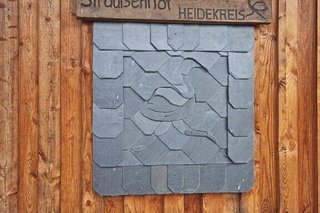 Ein aus Schieferplatten zusammengesetzter Strauß und ein Holzschild mit Hinweis Straußenfarm hängen an der Holzwand einer Scheune.