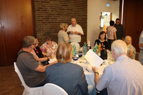 Mehrere Personen sitzen an einem runden Tisch. Dahinter stehen andere Personen und unterhalten sich.