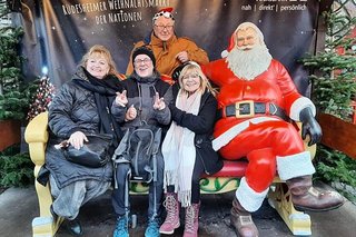 Zwei Frauen und ein Mann sitzen neben dem überlebensgroßen Weihnachtsmann aus Kunststoff auf einer rot-gold-angemalten Polsterbank, ebenfalls aus Kunststoff. Hinter ihnen hängt ein Banner mit Grüßen vom Rüdesheimer Weihnachtsmarkt der Nationen.