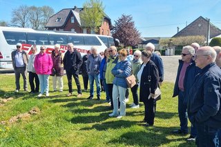 Eine Besuchergruppe steht auf einer Wiese und lässt sich erklären, wie Spargelanbau funktioniert. Im Hintergrund steht ein weißer Reisebus.