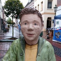 Skulptur zeigt Oberk&ouml;rper eines Jungen mit brauen Haaren, gr&uuml;ner offener Jacke und gelbem Pullover