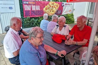 Eine Gruppe Menschen sitzt an einem Rastplatz-Tisch unter einem roten Langnese-Sonnenschirm. Im Hintergrund hängt ein Banner an einem Zaun mit der Aufschrift Gönn dir mal eine Pause.