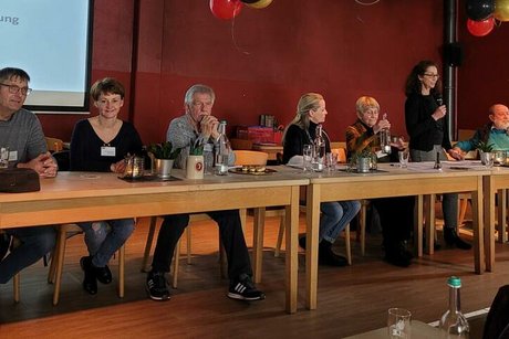 Drei Männer und vier Frauen sitzen gemeinsam an einer Tischreihe vor Kopf des Saales.