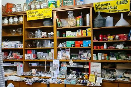 Allerhand Krimskrams und Drogerieartikel sind zwischen alten Apotheker-Glasflaschen und Blechdosen in einem alten Holzregal arrangiert.