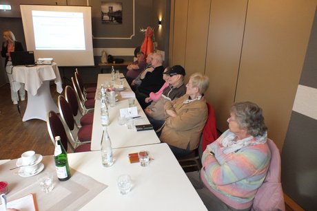 Mehrere Personen sitzen an langen Tischen und h&ouml;ren einem Vortrag zu.