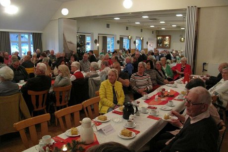 Viele Menschen sitzen in einem Saal an langen, weihnachtlich geschmückten Tischen.