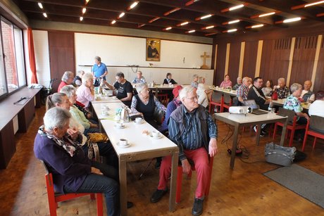 Mehrere Menschen sitzen an langen Tischen und h&ouml;re einem Referenten zu.