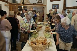 Menschen stehen um einen langen, weiß eingedeckten Tisch herum und bedienen sich an dem darauf aufgebauten Buffet.