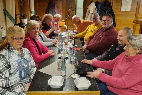 Eine Gruppe Männer und Frauen sitzt in einer Gaststube an einem langen Tisch beisammen.