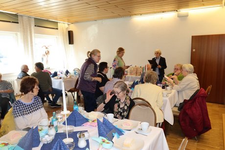 An mehreren gedeckten Tischen sitzen Menschen und unterhalten sich miteinander. Drei Frauen stehen dazwischen und sprechen mit den Menschen.
