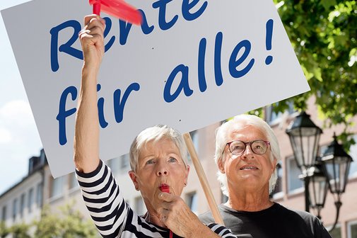 Seniorenpaar auf einer Renten-Demo Eine ältere Frau und ein älterer Mann mit einer Demopappe mit der Aufschrift "Rente für alle!". Die Frau bläst in eine Trillerpfeife, hält eine Ratsche in der Hand.