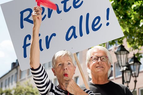 Eine ältere Frau und ein älterer Mann mit einer Demopappe mit der Aufschrift "Rente für alle!". Die Frau bläst in eine Trillerpfeife, hält eine Ratsche in der Hand. 