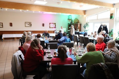 Frühlingsfest Mehrere Menschen sitzen an vier Tischen und hören dem Sprecher im Hintergrund zu.