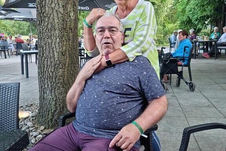 Eine Frau steht lächeln hinter einem Mann, der auf einem Bistrostuhl sitzt und streicht ihm über den Kopf.