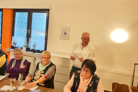 Drei Frauen sitzen an Tischen und ein Mann liest etwas von einem Zettel ab.