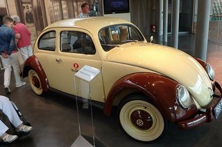 Ein auf Hochglanz polierter, beige-dunkelroter Käfer mit Dr. Oetker-Logo auf der Beifahrertür wird in der Ausstellung präsentiert. Daneben sind Besucher zu Fuß oder im Rollstuhl unterwegs.