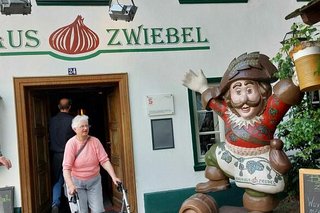 Eine Frau mit Rollator kommt gerade aus dem Brauhaus Zwiebel. Vom Betrachter aus rechts neben ihr steht die überlebensgroße Figur eines Mannes mit Dreispitz und Bierkrug in der Hand.