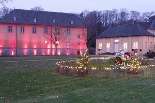 Ein Weihnachtsmann mit Schlitten ist als Dekoration hinter einem mit Lichterketten geschmückten Zaun aufgebaut. Im Hintergrund leuchtet ein rot angestrahltes Nebengebäude des Schlosses in der Dämmerung.