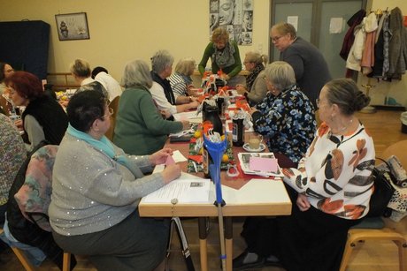 Jahresabschlusstreffen der Frauenvertreterinnen. Die Vertreterin der Frauen zeigt den am Tisch sitzenden Frauen, wie sie ein Blatt zu einer Figur faltet.