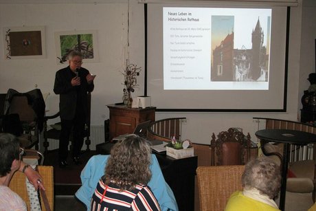 Viele Menschen sitzen gemeinsam in einem Raum an Tischen und hören einem Mann zu, der mit Mikrofon vor einer PowerPoint-Präsentation steht.