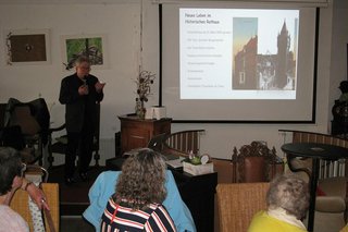 Viele Menschen sitzen gemeinsam in einem Raum an Tischen und hören einem Mann zu, der mit Mikrofon vor einer PowerPoint-Präsentation steht.