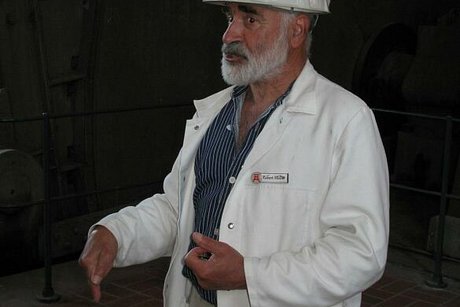 Ein bärtiger Mann mit weißem Bauhelm und Bergmannsjacke steht gestikulierend in einer dunklen Halle.