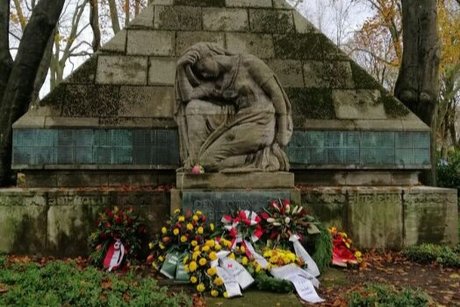 Blumenkränze liegen vor dem steinernen Ehrenmal, dass eine kniende Frau vor einer Pyramide aus Blocksteinen darstellt.