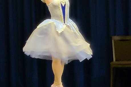 Eine junge Frau in weißem Ballettkostüm und Spitzenschuhen steht in Tanzpositur auf der Bühne.