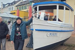 Ein Mann in dunklem Weihnachtspulli und blauer Steppjacke posiert neben einem an Land aufgestellten kleinen Kutter namens "Purzel"