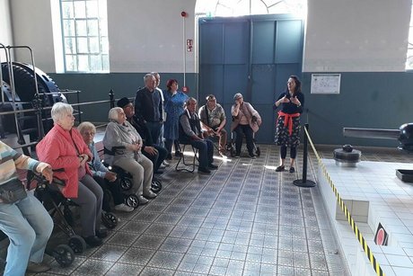 Eine Gruppe Menschen steht oder sitzt auf Rollatoren in einer großen, gefliesten Halle und hört einer Frau zu, die etwas erzählt.
