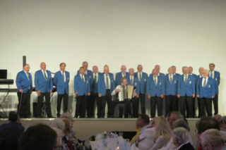 Männer in blauen Sakkos stehen singend auf der Bühne, in der Mitte ein Mann mit Akkordeon.