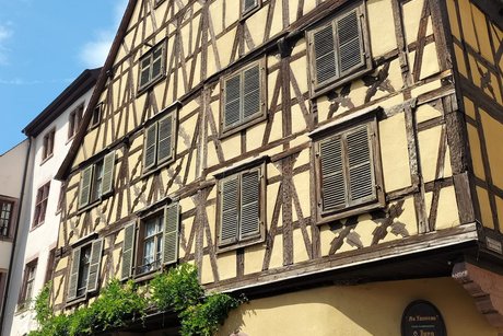 Zu sehen ist ein Eckhaus; im Erdgeschoß ein Weingeschäft mit Passanten vor den Fenstern; die oberen Etagen zeigen den Reichtum der Erbauer mit wunderschönem Fachwerk.