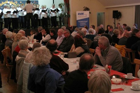 Viele Menschen sitzen in einem Saal an langen, weihnachtlich geschmückten Tischen. Im Hintergrund befindet sich eine Bühne, auf der gerade ein Chor singt.