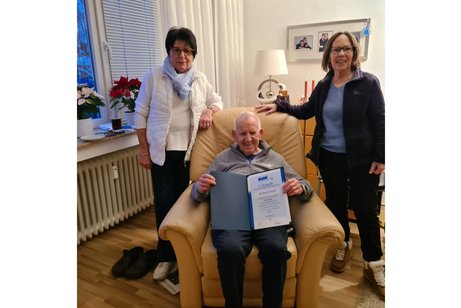 Zwei Frauen stehen rechts und links neben einem Sessel, in dem ein Mann sitzt.