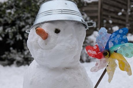 Ein Schneemann mit Möhrennase, Metallschüsselhut und buntem Windrad vor verschneiter Landschaft.