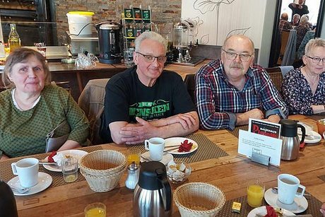 Zwei Frauen und zwei Männer sitzen gemeinsam am gedeckten Café-Frühstückstisch und schauen in die Kamera..