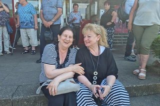 Zwei Frauen sitzen gemeinsam auf einer breiten Treppe aus Stein. Im Hintergrund stehen und laufen weitere Menschen umher.