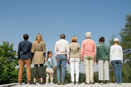 Menschen verschiedenen Alters mit und ohne Behinderung stehen nebeneinander, wenden dem Betrachter den R&uuml;cken zu. Eine kleinw&uuml;chsige junge Frau dreht sich zum Betrachter um.