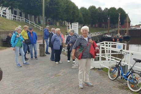 Eine Gruppe Menschen steht am Ufer eines Kanals. Im Hintergrund sieht man eine Allee und einige Schiffe, die am Ufer vertäut sind.