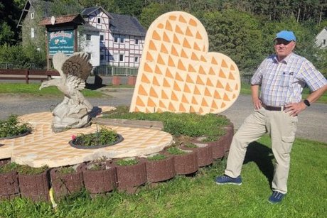 Ein Mann mit Schirmmütze posiert neben der Skulptur aus Herzwaffelstücken und steinernem Greifvogel, das in einem Rondell vor dem Waffelhaus aufgestellt ist.