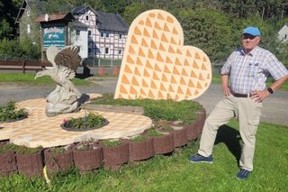 Ein Mann mit Schirmmütze posiert neben der Skulptur aus Herzwaffelstücken und steinernem Greifvogel, das in einem Rondell vor dem Waffelhaus aufgestellt ist.