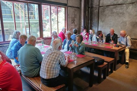 Menschen sitzen gemeinsam an Tischen vor einer großen Fensterfront.