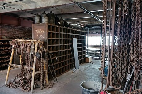 Eine ehemalige Werkstatthalle der Kohlenwäscherei mit Regalen und rostigen Metallketten, die von der Decke hängen.