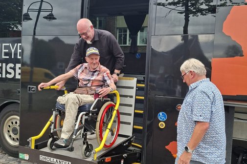 Im Hintergrund ein Bus auch f&uuml;r Rollstuhlfahrer geeignet. Es wird ein Rollstuhl in den Bus mit einer Hebeb&uuml;hne bef&ouml;rdert.